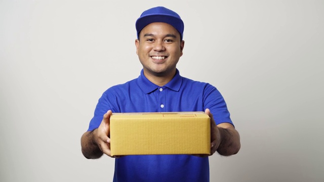 微笑年轻的亚洲快递员在蓝色制服给包裹硬纸板客户在孤立的白色背景。4 k决议。视频下载