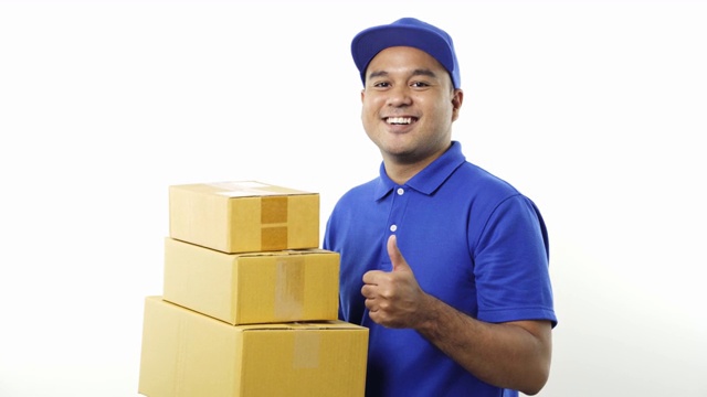 微笑的年轻亚洲快递员在蓝色制服显示大拇指举着包裹纸箱在孤立的白色背景。4 k决议。视频下载
