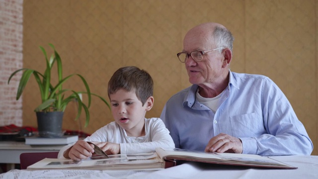 快乐的时刻，快乐的小男孩和他快乐的爷爷一起回忆着家人，坐在房间的桌子旁看着相册视频下载
