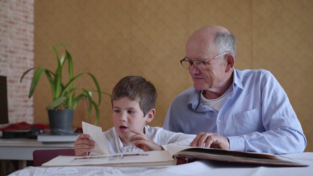 家人的回忆，小快乐的孩子和他快乐的爷爷，坐在桌子前看相册的房间视频下载