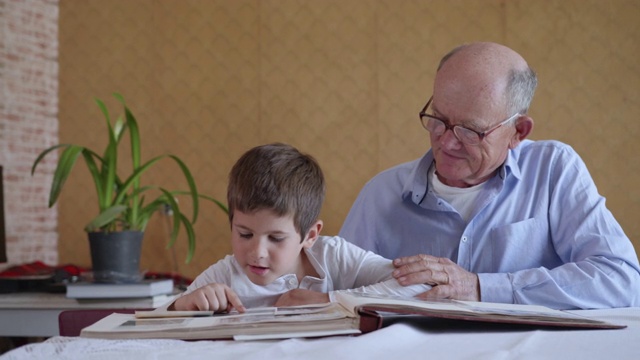 戴着眼镜的老爷爷和好奇的小男孩在一起，享受着快乐的回忆，讲述着过去的岁月，坐在房间的桌子前看着家庭相册视频下载