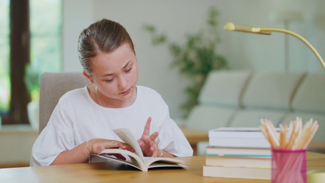 一个专注的少女坐在书桌前，一边看书一边翻着书。书、笔记本、铅笔、笔记本、台灯对第一个计划失去焦点。背景是模糊的视频素材