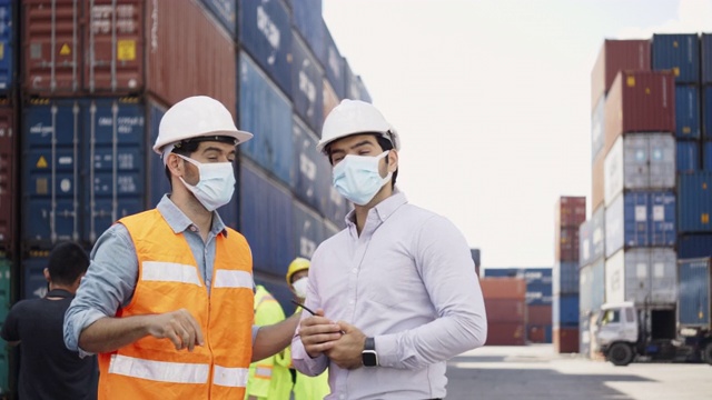 集团工程师和施工人员在工作中戴医用口罩和防护设备如安全帽。团队合作在建筑工业工厂有集装箱柜运输。概念的工作视频素材