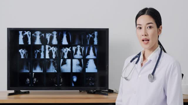 一名身穿实验服的亚洲女性，一边对着摄像机，一边在屏幕上解释x光胶片。视频下载