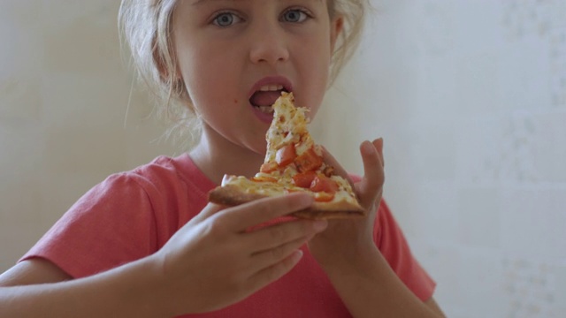 孩子吃披萨。饥饿的小女孩坐在厨房的餐桌上吃着美味的意大利披萨。不健康的营养快餐。一个吃午饭的孩子的肖像。面对年轻女孩特写。视频素材