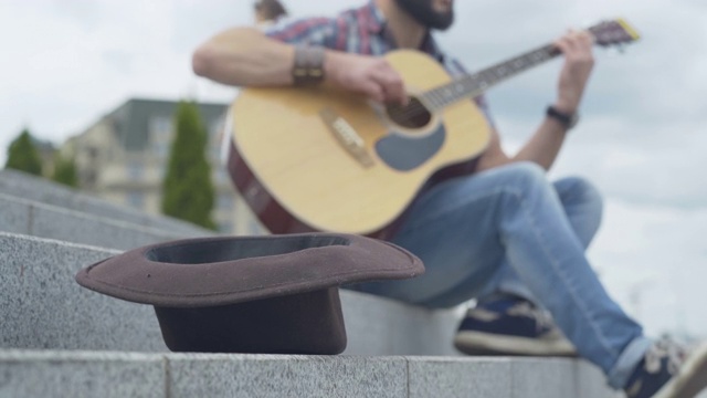 城市楼梯上棕色帽子的特写，背景是一个模糊的男人在弹吉他。一个不知名的白人吉他手在城市在夏天的一天表演。视频素材