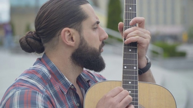 一个人在户外弹吉他的侧视图。在城市街道上表演的留着胡子的年轻白人音乐家的肖像。有创意的家伙在用乐器。视频素材