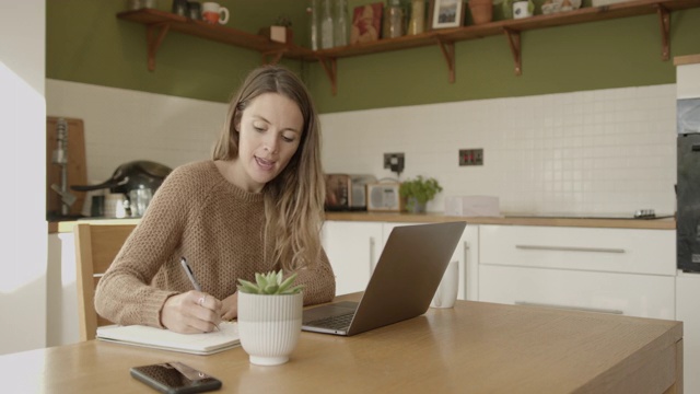 女性在和同事视频通话，在厨房的桌子上用笔记本电脑工作视频素材