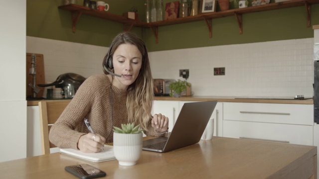 女性在视频通话中使用耳机，在流感大流行期间在家厨房餐桌旁工作视频素材