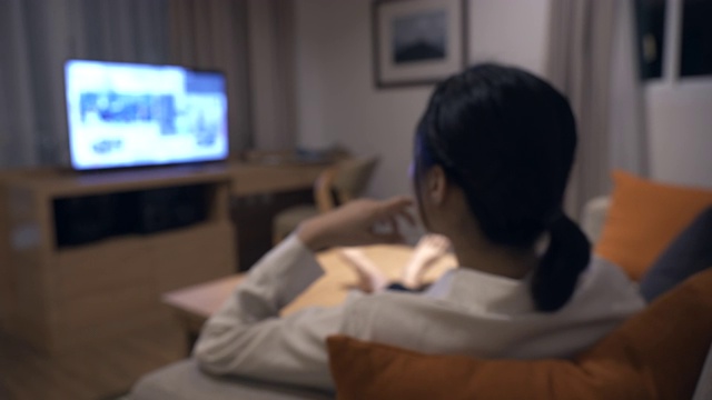 一个亚洲女人坐在沙发上看电视的场景视频下载