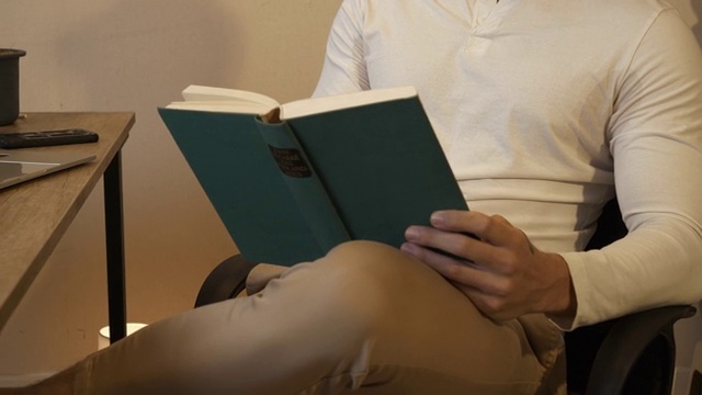 男性手握书的特写视图。这个年轻人坐在家里的椅子上看书。视频素材