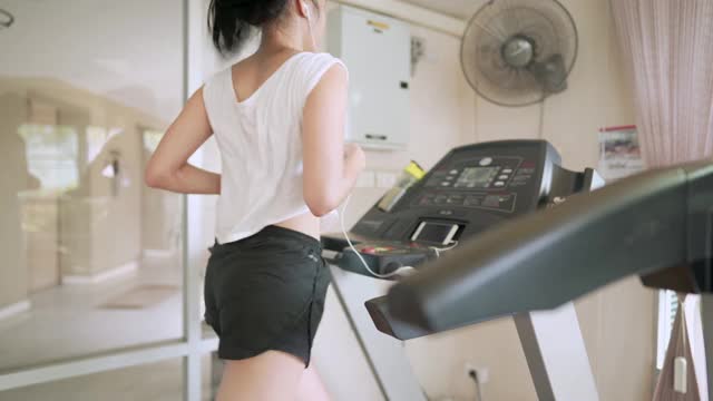 亚洲年轻女性在健身房的跑步机上跑步，一边听音乐一边做有氧运动，进行减重耐力训练，现代活跃的生活视频下载