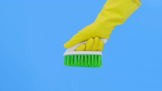 一只手戴着黄色手套，在蓝色背景下显示绿色刷子，清洁和刷地毯，去除污渍和羊毛，做日常作业概念视频下载
