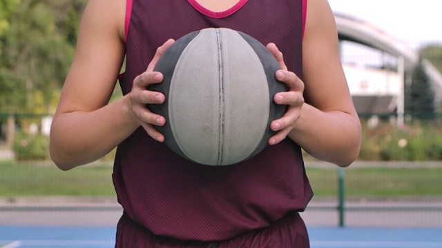 一个篮球运动员手里拿着一个球，对着镜头。夏天进行户外训练。健康和积极的生活方式。球和人的躯干特写。慢动作视频下载