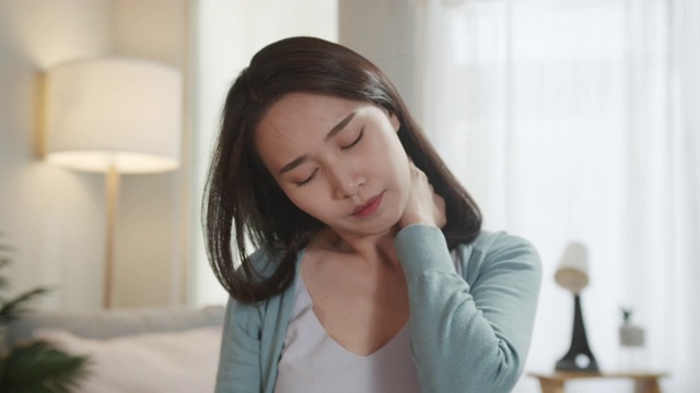 疲惫的亚洲女人坐在客厅里，感觉疲惫，脖子疼痛，肩膀被碰，脸上疼痛。保健和身体问题的概念。视频素材