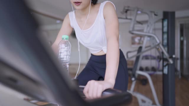 活跃的年轻亚洲女人戴着耳机听着音乐享受自行车运动在健身房快乐快乐的自行车，口渴的运动女孩喝着水，有氧燃烧脂肪的训练计划，自然的白天光线视频素材