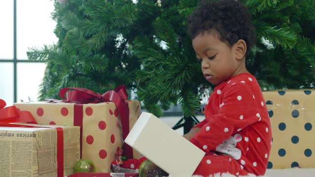 快乐的婴儿男孩非洲民族年龄12个月打开圣诞礼盒，同时把礼物放在圣诞树下。亲密的圣诞节的概念。视频下载