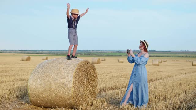 暑假，年轻快乐的妈妈用复古相机拍下了收获后兴高采烈的儿子在田野的草堆上的照片视频下载
