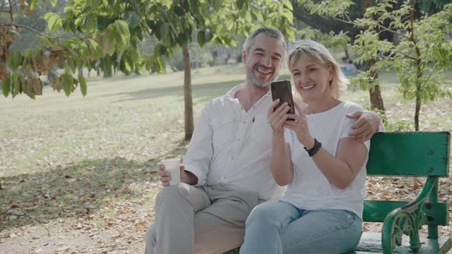 老夫妇在公园里玩智能手机。家人带着幸福的微笑，在清晨与自然放松。或者在晚上。进入老年社会，退休。概念医疗保险视频素材