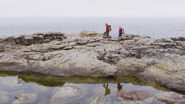 一对老夫妇背着背包在海边的岩石上徒步旅行。视频下载