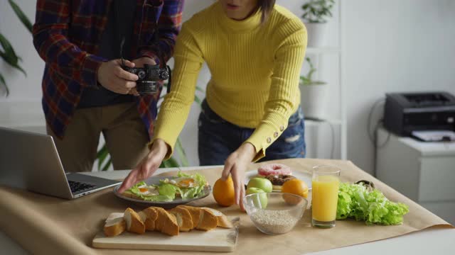 女性平面造型师在桌子上布置物品，帮助男性摄影师拍摄食物与水果和食物的背景照片视频下载