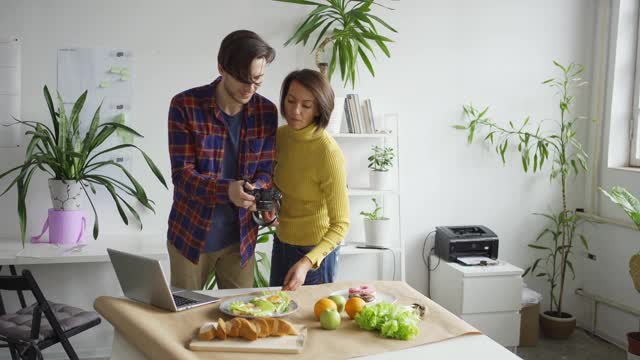 女性平面造型师帮助男性摄影师拍摄食物背景照片与水果和食物放在办公室的桌子上视频下载