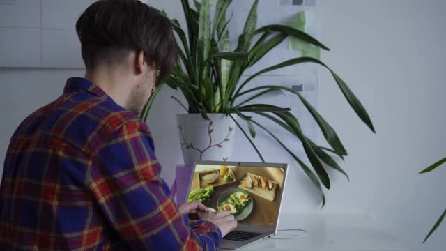 放大镜头，年轻的摄影师在笔记本电脑上查看图片，享受在工作室桌子上放食物的平躺照片视频下载