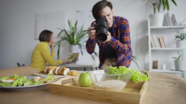 男摄影师拍摄餐桌上食物的特写跟踪镜头，女制片人在她的帮助下查看笔记本电脑上的照片并给出建议视频下载
