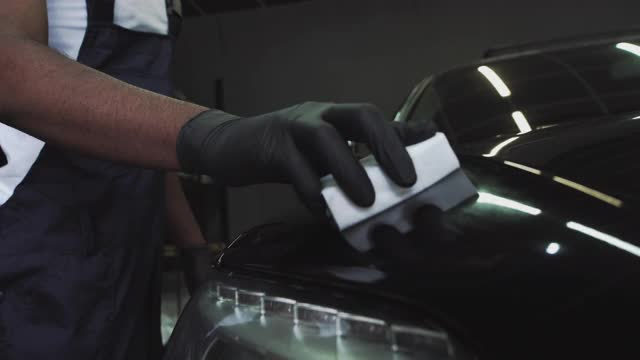 一名非裔美国服务人员涂抹了一层保护层。用于保护车灯。专业汽车陶瓷工在车窗上涂上一层陶瓷防护雨罩。概念来自纳米保护，不同的配件，长视频素材