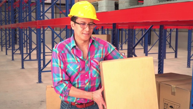 在一个空而干净的配送仓库里，一名资深亚洲工人面带微笑，情绪积极，戴着一顶黄色的防护帽，将一个已递送的纸板箱作为镜头的视点。视频下载