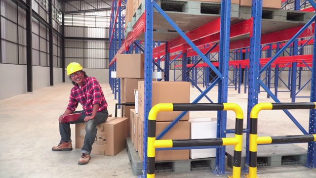一位资深亚洲工人微笑着，情绪积极，头上戴着一顶保护帽，坐在一个纸箱上。这个纸箱是一名邮政工人将包裹送到一个空的配送仓库的。视频下载
