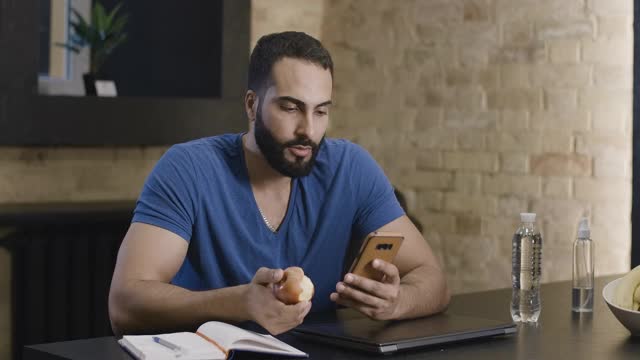 年轻英俊的中东男子，一边用智能手机上网一边吃苹果。快乐自信的男人使用社交媒体或在线消息的肖像。电影4k ProRes总部。视频素材
