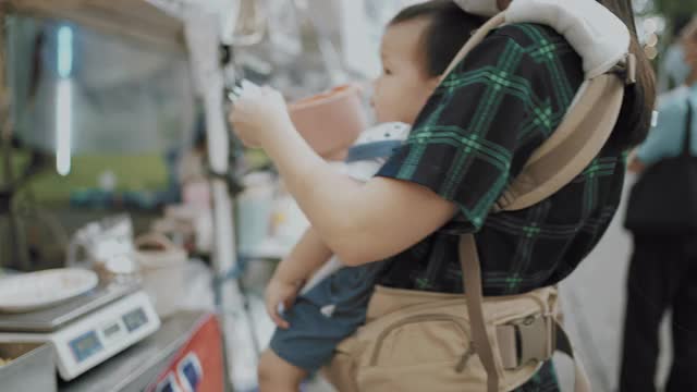 年轻的亚洲母亲抱着她的儿子购物和享受在街头食物视频下载