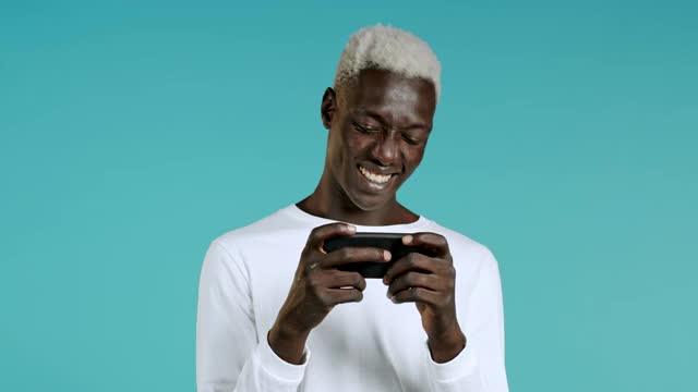 英俊的非洲男人在工作室的蓝色墙上玩智能手机游戏。使用现代技术，应用程序，社交网络。视频素材