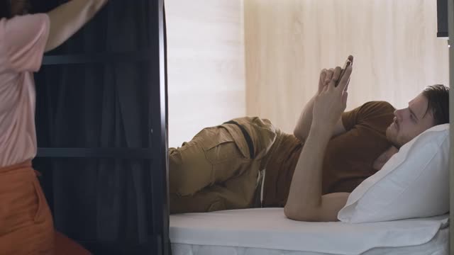 照片中，一名男子躺在旅馆的床上使用智能手机，而另一名女子正在爬梯子。年轻的白种人睡在旅店里。电影4k ProRes总部。视频素材