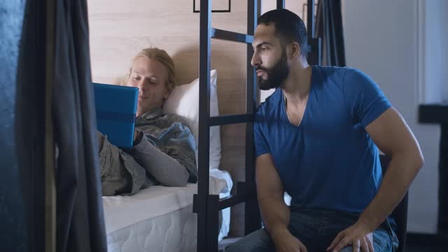 英俊的中东男子与轻松的白人朋友聊天躺在床上与笔记本电脑。无忧无虑的多民族人在晚上的青年旅社或客栈。电影4k ProRes总部。视频素材