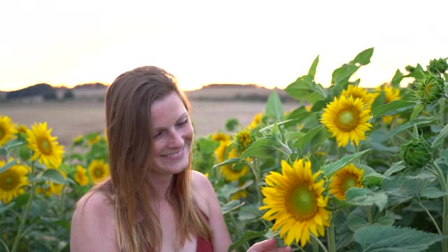 在阳光明媚的日子里，一个金发碧眼的白人女人在农场里闻着向日葵的香味。高质量4k镜头视频素材