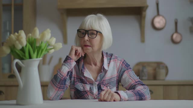 精力充沛的70多岁老妇人成功举起双手，坐在白色郁金香的餐桌上。快乐的金发老女人在家。视频素材
