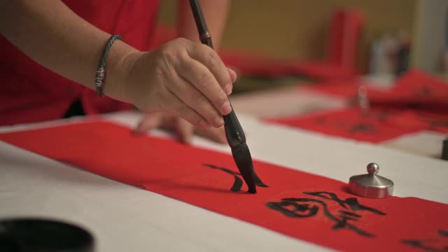 亚洲华人男性练习书法的手，为即将到来的中国新年庆祝家庭装饰目的繁荣和良好的措词写在一张红色的纸上视频下载