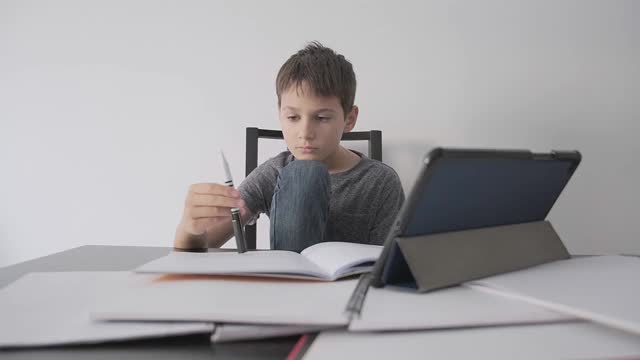 悲伤的孩子坐在写字台上，拿着平板电脑和很多书。男孩感到很无聊，因为他看不懂作业，他玩笔。教育，学习困难，在线学习理念。视频素材