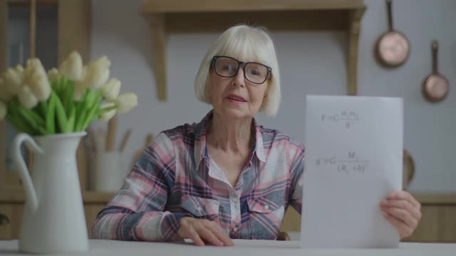 戴着眼镜的高级教师在镜头前展示一张纸，上面写着物理方程。在线教育和远程学习过程。网络摄像头的观点，在家工作的妇女交谈。视频素材
