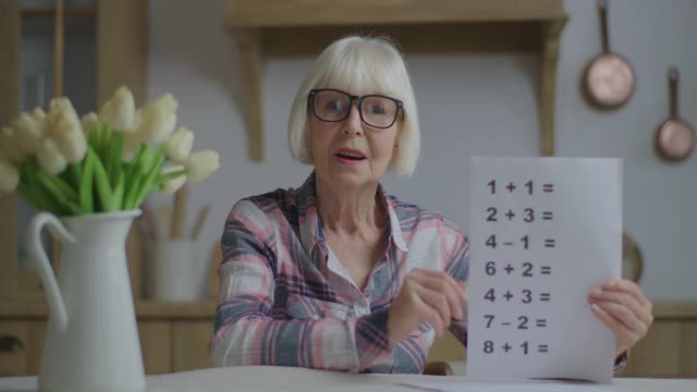 戴着眼镜的高级教师在镜头前展示一张纸，上面有简单的数学方程。在线教育和远程学习过程。网络摄像头的观点，在家工作的妇女交谈。视频素材