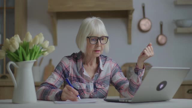 高级教师戴着眼镜用钢笔在纸上写字看着笔记本电脑。在线教育和远程学习过程。在家工作的妇女签署文件。视频素材