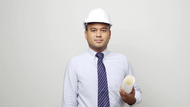 年轻英俊的亚洲土木工程师拿着图纸卷站在工作室里。工人在孤立的白色背景上戴着安全帽。4 k决议。视频下载