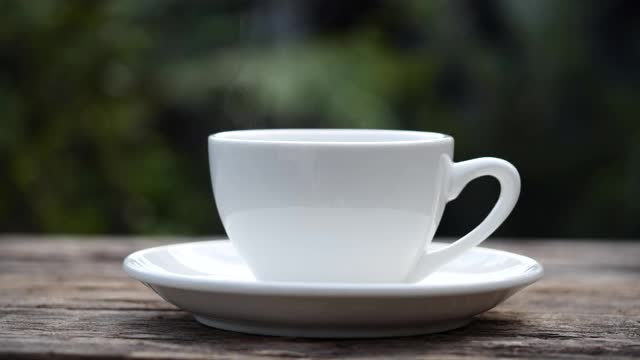 特写热气腾腾的咖啡或茶杯，慢动作。热陶瓷白色咖啡杯与烟在旧木桌上的自然背景。视频素材
