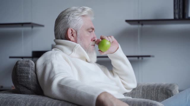 满脸胡须的老人独自坐在椅子上，手里拿着一个苹果，思考着视频素材