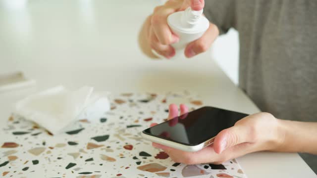 一名女子用手在手机屏幕上喷洒消毒剂，并用纸巾擦拭视频素材