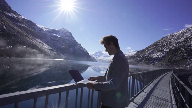 一名身穿灰色西装的男子在一个湖泊和雪山附近的大坝上使用笔记本电脑和手机视频下载