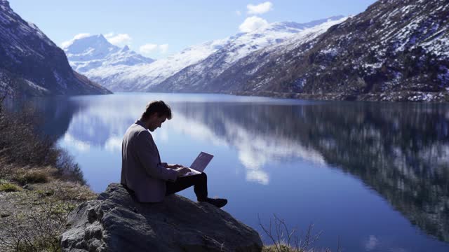 一名身穿灰色西装的男子在湖边和雪山边使用笔记本电脑视频下载