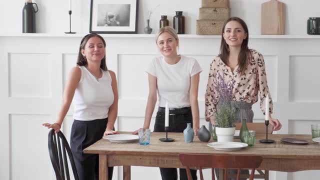 三个女孩看着相机，微笑着站在厨房里，摆好盘子准备一起吃晚餐视频下载